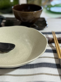 Ceramic bowl with a spoon on a striped tablecloth, with chopsticks and additional bowls in the background.