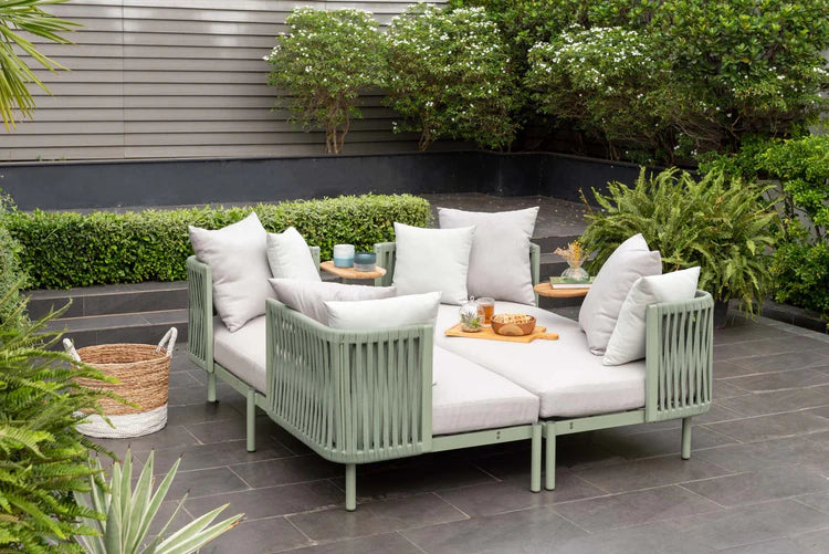 Outdoor patio set with green frames and white cushions on a stone patio, surrounded by plants.