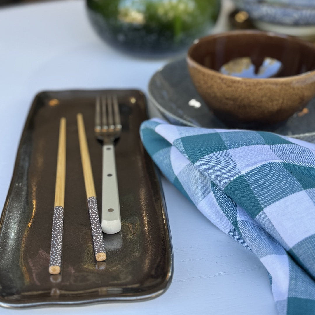 Ceramic tray with chopsticks and a fork, green and blue checkered napkin, and a blurred background.