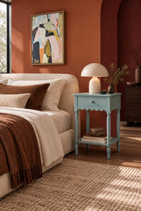 A styled bedroom scene featuring a light blue nightstand beside a cream bed, with a white table lamp, a framed abstract painting on a terracotta wall, and warm natural light.