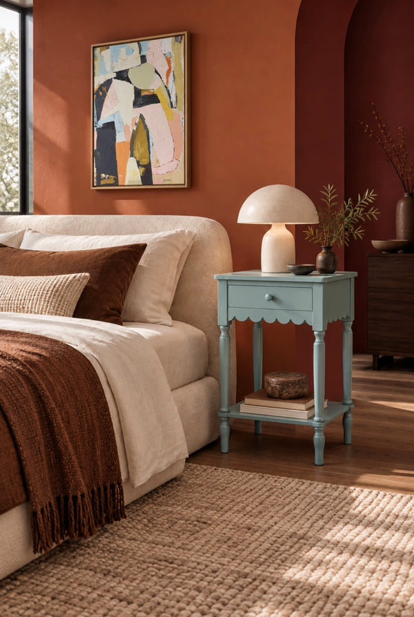 A styled bedroom scene featuring a light blue nightstand beside a cream bed, with a white table lamp, a framed abstract painting on a terracotta wall, and warm natural light.