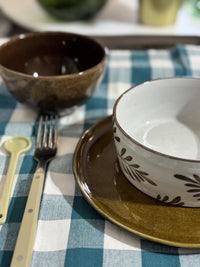 Dinnerware set on a checkered tablecloth with a brown bowl, white bowl, and gold fork.