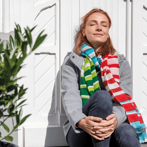 Woman wearing colorful striped scarves sitting against a white wooden wall.