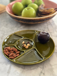 Green ceramic plate with compartments containing nuts and a cherry, placed on a marble surface with apples in the background.