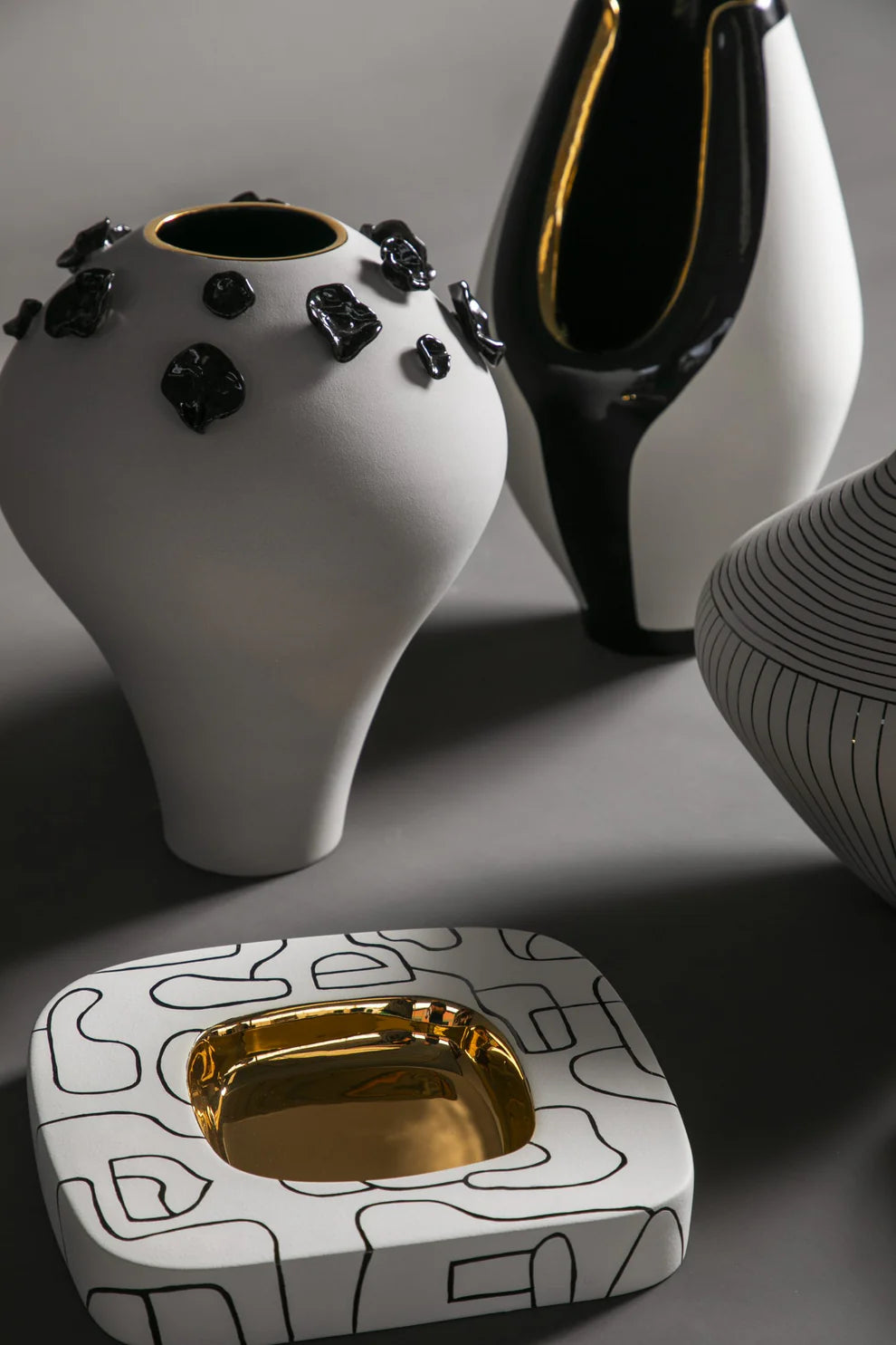 Decorative ceramic items including a white vase with black accents, a black and gold pitcher, and a square dish with gold interior on a gray background.