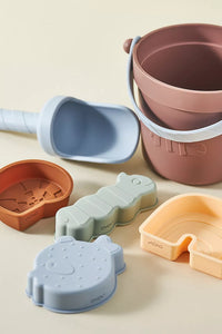 Set of colorful silicone sand toys including a bucket, shovel, and molds on a beige background.