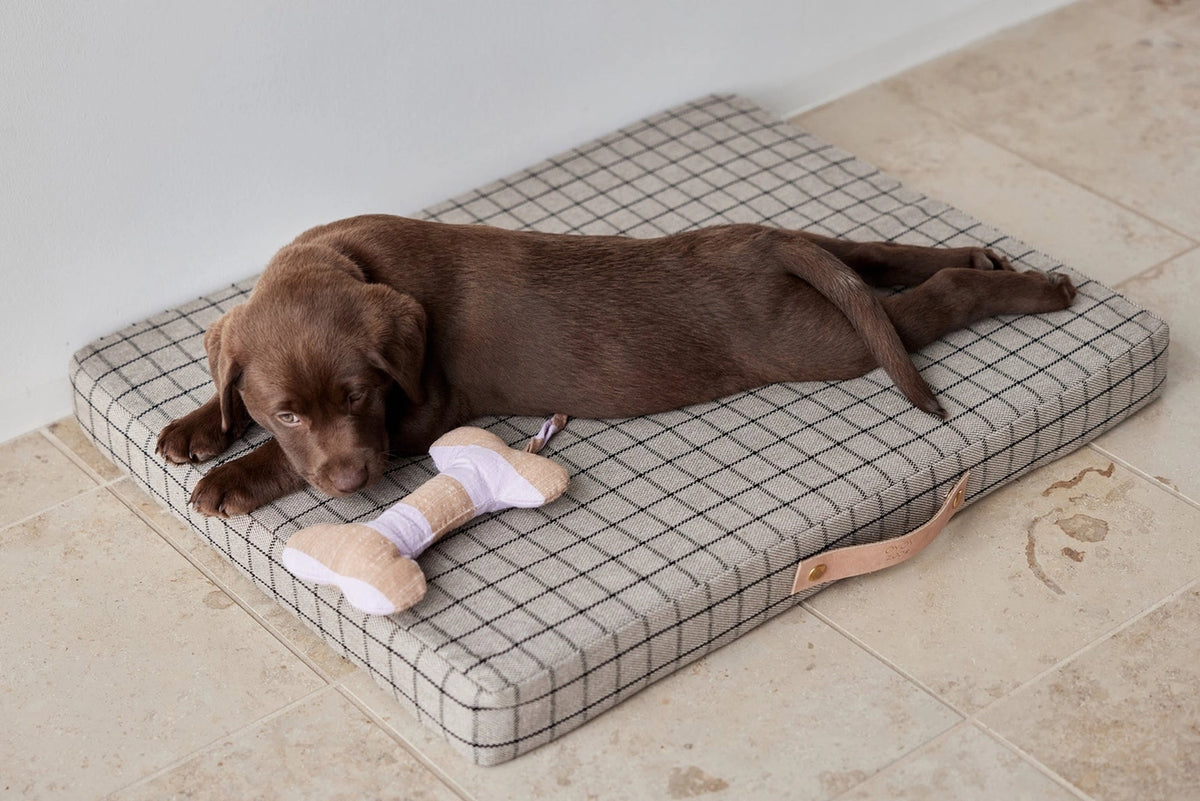 A brown puppy lies on a square cushioned pet bed with a gray and white grid pattern, next to a small bone-shaped toy, on a beige tiled floor.