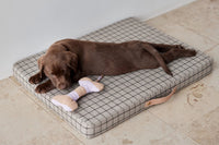 A brown puppy lies on a square cushioned pet bed with a gray and white grid pattern, next to a small bone-shaped toy, on a beige tiled floor.
