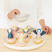 Decorative cookies shaped like people on a plate with a person holding a bowl in the background.