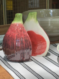 Two ceramic condiment shakers shaped like figs on a striped tablecloth.