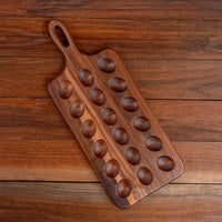 A rectangular wooden serving board with a loop handle and ten oval recessed bowls, photographed from above on a rustic wooden tabletop.