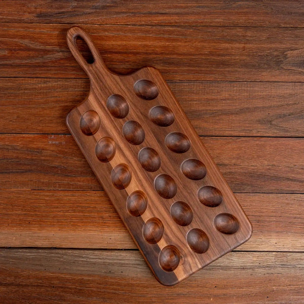 A rectangular wooden serving board with a loop handle and ten oval recessed bowls, photographed from above on a rustic wooden tabletop.