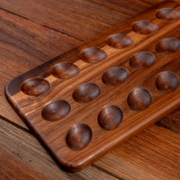 A rectangular wooden board with 16 carved circular wells, photographed at an angle on a matching wooden surface.