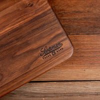 A close-up of a dark brown wooden cutting board with rounded corners and a laser-engraved 'Scotman Trade Co Mark' logo, resting on a rustic wooden plank surface.