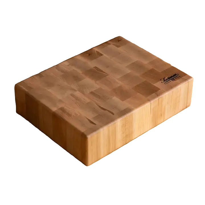 A rectangular wooden cutting board with a visible grain pattern and a small black brand logo in the top right corner, resting on a wooden countertop with a black tiled backsplash behind it.