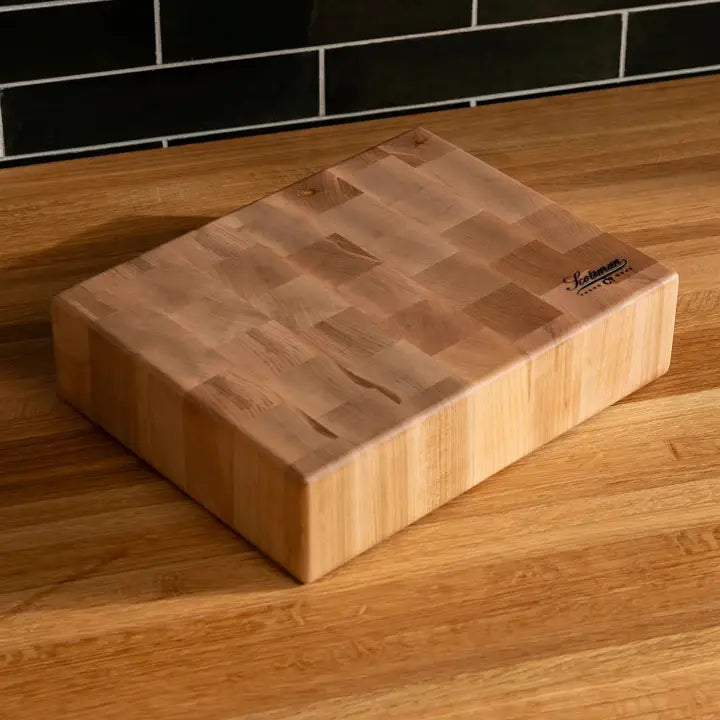 A rectangular wooden cutting board with a visible grain pattern and a small black brand logo in the top right corner, resting on a wooden countertop with a black tiled backsplash behind it.