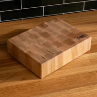 A rectangular wooden cutting board with a visible grain pattern and a small black brand logo in the top right corner, resting on a wooden countertop with a black tiled backsplash behind it.