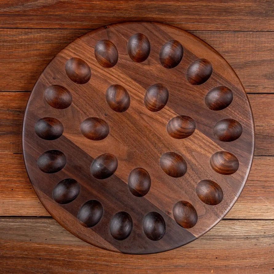 A round wooden board with 24 carved oval pieces arranged in a circular pattern, set on a rustic wooden plank background.
