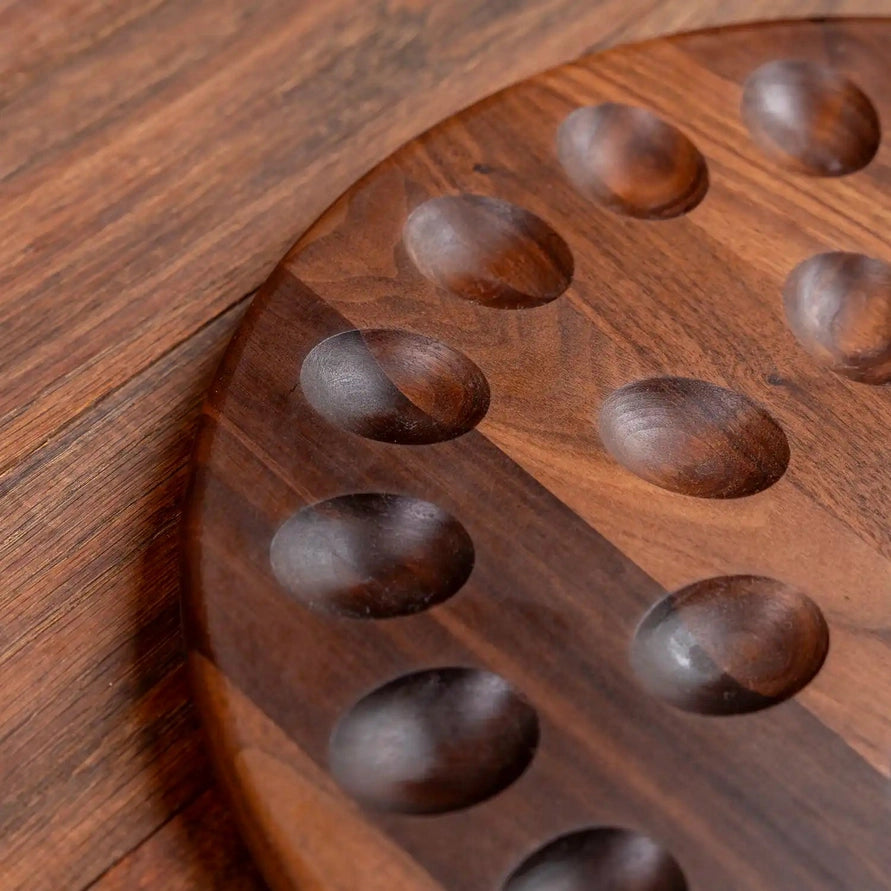A close-up of a round wooden board with carved oval indentations, photographed on a matching wood surface.