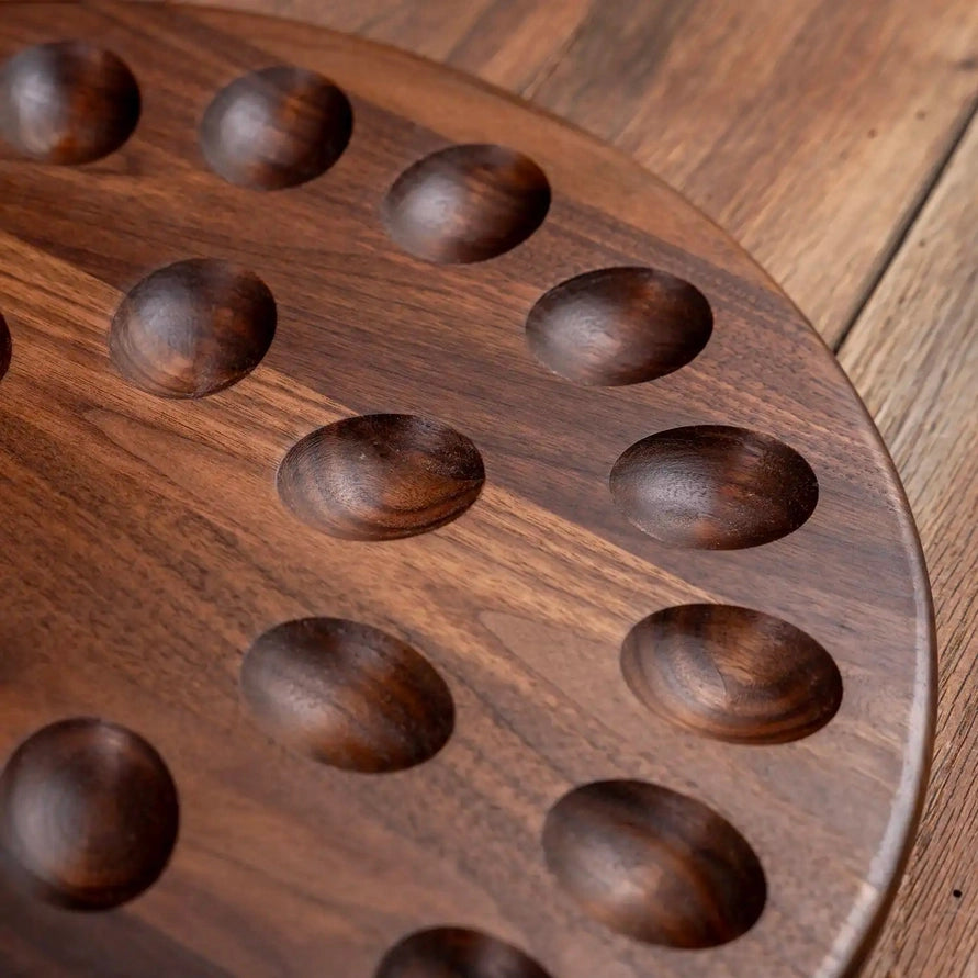 Close-up of a round dark brown wooden board with multiple oval recessed pockets, photographed on a matching wood surface.