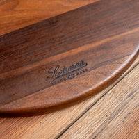 A close-up of a dark brown wooden board with rounded corners and a debossed 'Shotman Made Co. Marr' logo, resting on a light brown wooden surface.