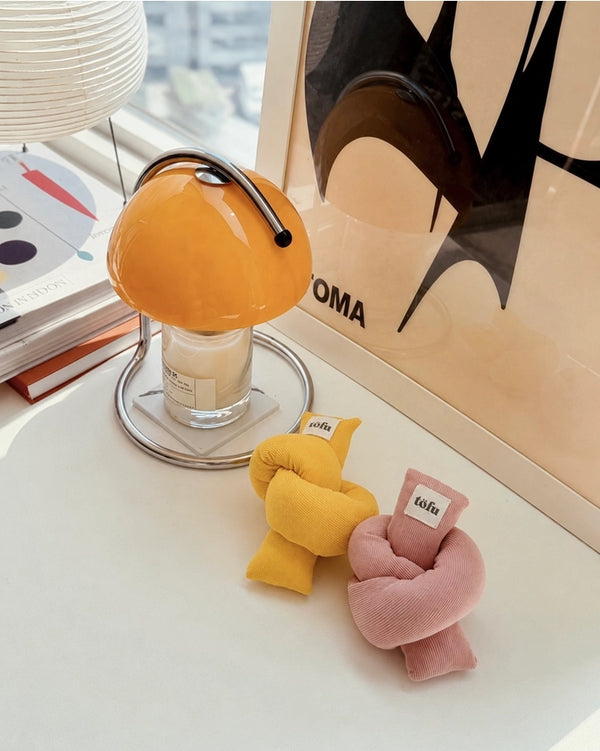 Two small plush decorative pillows in yellow and pink, each with a small white tag reading 'tofu', arranged on a white tabletop beside a mushroom-shaped glass candle holder and a framed abstract print.