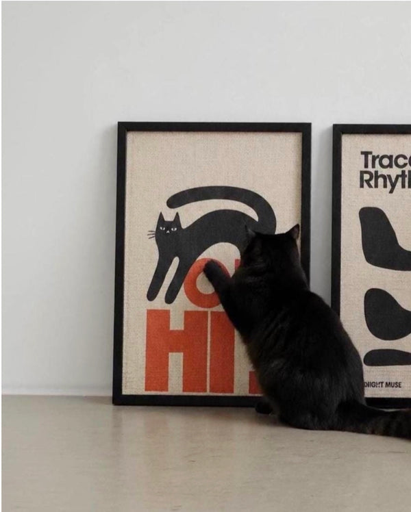 A black cat sits on a light floor, touching the corner of a framed art print. The print features a minimalist black cat silhouette over bold orange and beige lettering.