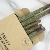 A cardboard box of catnip stick snacks, showing several green-brown dried herb sticks partially pulled from the box on a white marble surface.