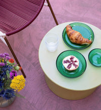 green stoneware side table on a purple floor with 3 green glass plates with fruit and a croissant