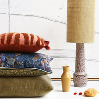 stack of 3 pillows next to a table lamp. Top pillow is red, mifddle pillow has a blue flower pattern and bottom pillow is green velvet