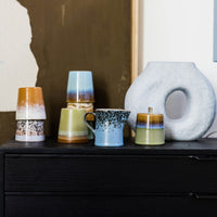 black dresser with brown, blue, and green colored mugs and a very light blue, organic shape vase object